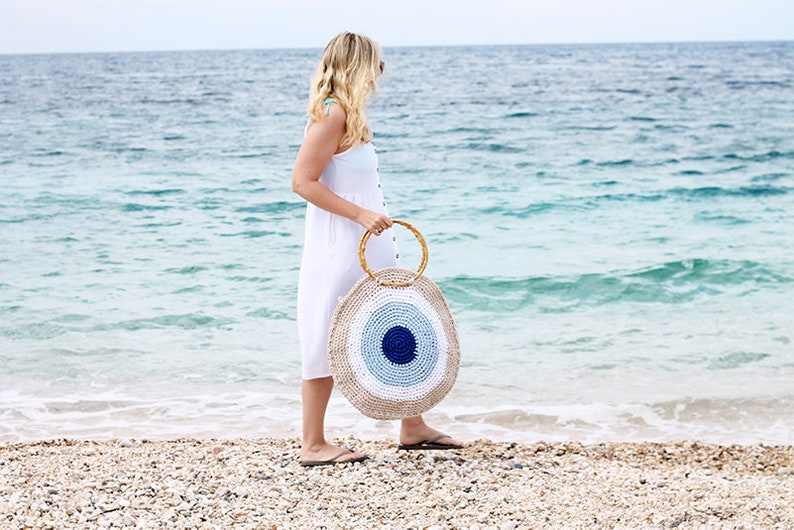 May include: A round, woven beach bag with a bamboo handle. The bag features a blue and white eye design. The person in the image is wearing a white dress and standing on a beach with the ocean in the background.
