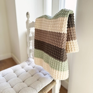 May include: A handmade crochet blanket draped over a white chair. The blanket features horizontal stripes in cream, brown, and green. The chair has a woven seat with a light gray cushion. The blanket's texture is visible.