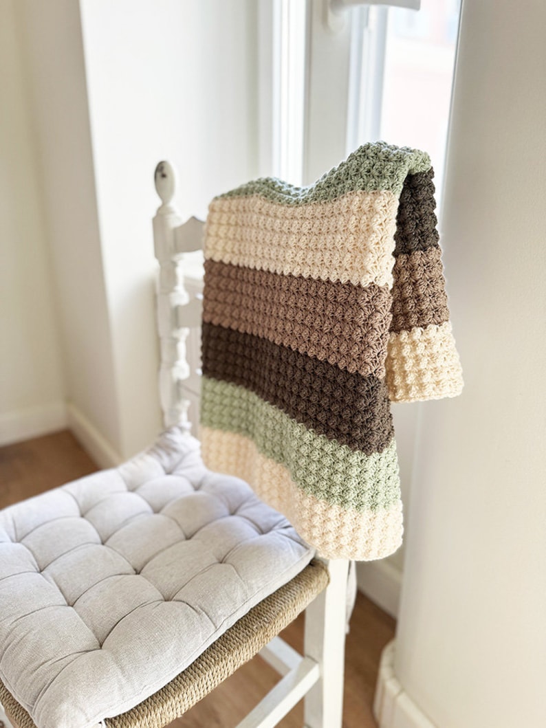 May include: A handmade crochet blanket draped over a white chair. The blanket features horizontal stripes in cream, brown, and green. The chair has a woven seat with a light gray cushion. The blanket's texture is visible.