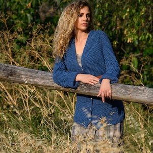 K&ouml;nnte beinhalten: Eine Frau mit langen, lockigen blonden Haaren tr&auml;gt einen blauen Strickcardigan mit Kn&ouml;pfen. Sie steht in einem Feld mit hohem Gras und lehnt sich an einen Holzlattenzaun.