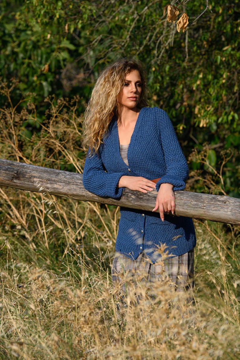 K&ouml;nnte beinhalten: Eine Frau mit langen, lockigen blonden Haaren tr&auml;gt einen blauen Strickcardigan mit Kn&ouml;pfen. Sie steht in einem Feld mit hohem Gras und lehnt sich an einen Holzlattenzaun.