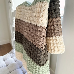 May include: A handmade, textured crochet blanket in a neutral color palette. The blanket features alternating stripes of cream, sage green, brown, and dark brown. The blanket is draped over a white wooden structure, showcasing its soft texture and design.