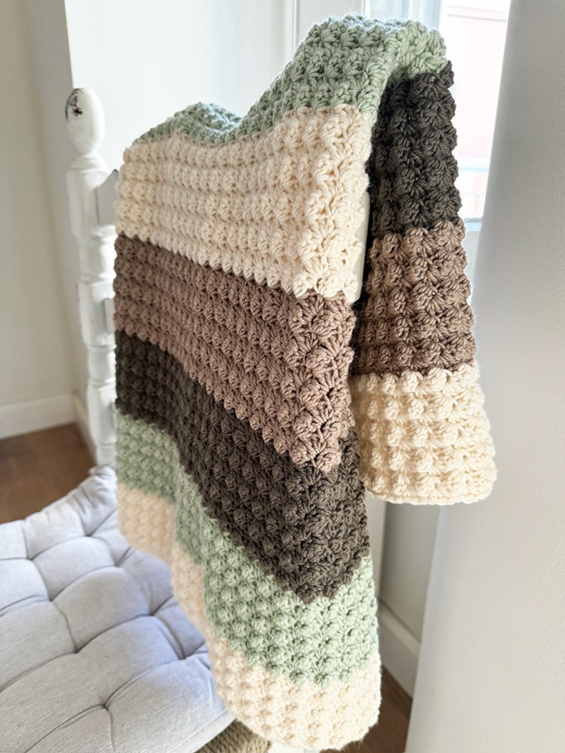 May include: A handmade, textured crochet blanket in a neutral color palette. The blanket features alternating stripes of cream, sage green, brown, and dark brown. The blanket is draped over a white wooden structure, showcasing its soft texture and design.