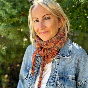 May include: A woman with blonde hair is wearing a denim jacket and a colorful knitted scarf. The scarf is a mix of orange, pink, and purple stripes.