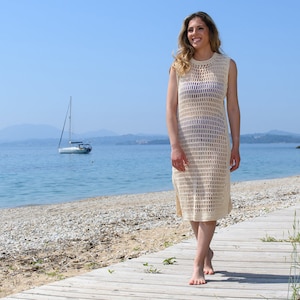 May include: A woman wearing a white crochet beach cover-up dress with a sleeveless design. She is walking on a wooden boardwalk near a beach.
