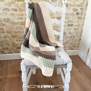 May include: A handmade, textured blanket in a neutral color palette of cream, sage green, brown, and gray, draped over a distressed white chair. The blanket features a woven pattern and adds a cozy touch to the setting.