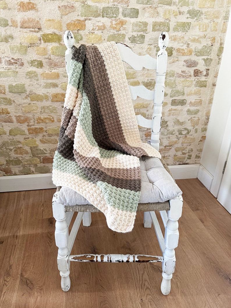 May include: A handmade, textured blanket in a neutral color palette of cream, sage green, brown, and gray, draped over a distressed white chair. The blanket features a woven pattern and adds a cozy touch to the setting.