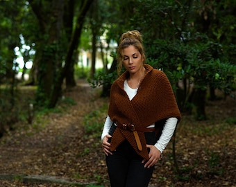 May include: A woman wearing a brown knitted shawl with a belt, over a white long-sleeved shirt and black pants. The shawl is wrapped around the shoulders and cinched at the waist with a brown leather belt. The background is a blurred forest.