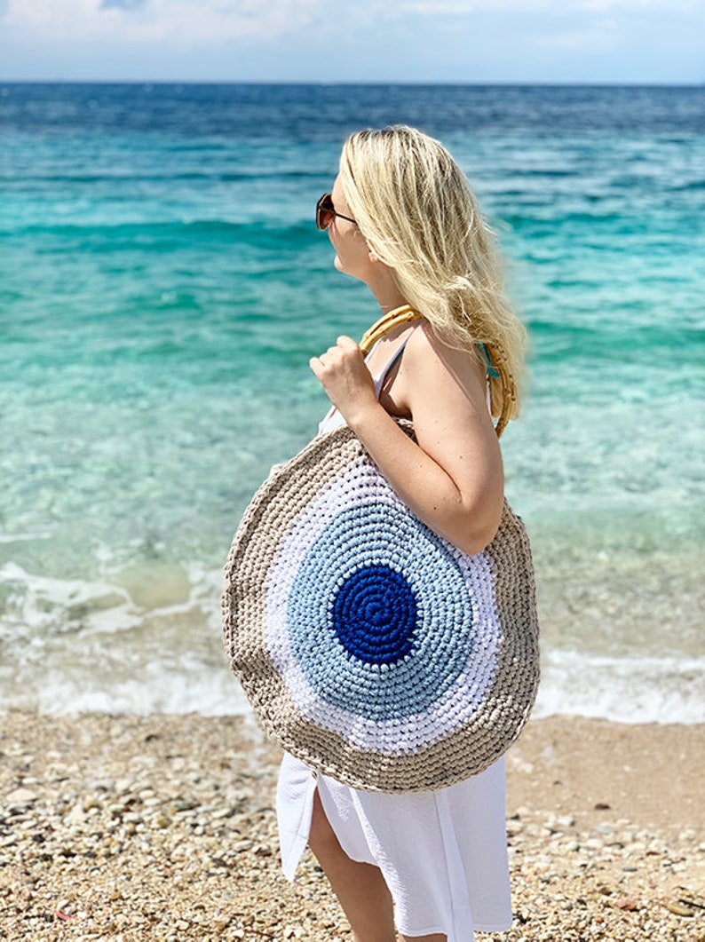 May include: A round, woven beach bag with a blue, white, and tan eye design. The bag has a dark blue center, surrounded by concentric circles of light blue, white, and tan. It has bamboo handles and is carried on a beach.