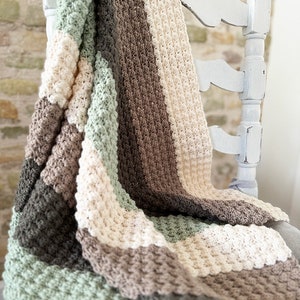 May include: A handmade crochet blanket draped over a white distressed chair. The blanket features a textured stitch pattern in alternating stripes of gray, sage green, cream, and brown. The cozy, soft blanket is perfect for home decor.