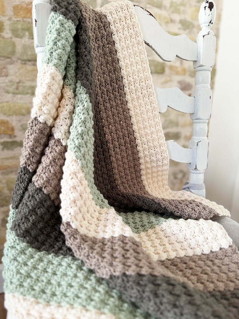 May include: A handmade crochet blanket draped over a white distressed chair. The blanket features a textured stitch pattern in alternating stripes of gray, sage green, cream, and brown. The cozy, soft blanket is perfect for home decor.
