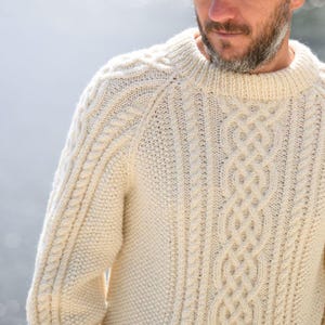 Puede incluir: Suéter de punto trenzado color crema con cuello redondo. El suéter presenta intrincados patrones de trenzas en la parte delantera y las mangas, con un diseño de punto texturizado. La prenda de punto gruesa y cálida es adecuada para el clima frío.
