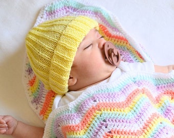 May include: A baby wearing a yellow knitted hat and a brown pacifier, sleeping on a rainbow crochet blanket.