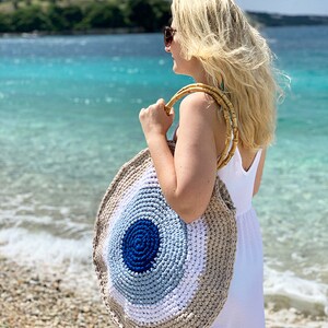 May include: A round, crocheted beach bag with a bamboo handle. The bag features concentric circles of color: dark blue, light blue, white, and beige. The bag is being held on a beach with turquoise water in the background.