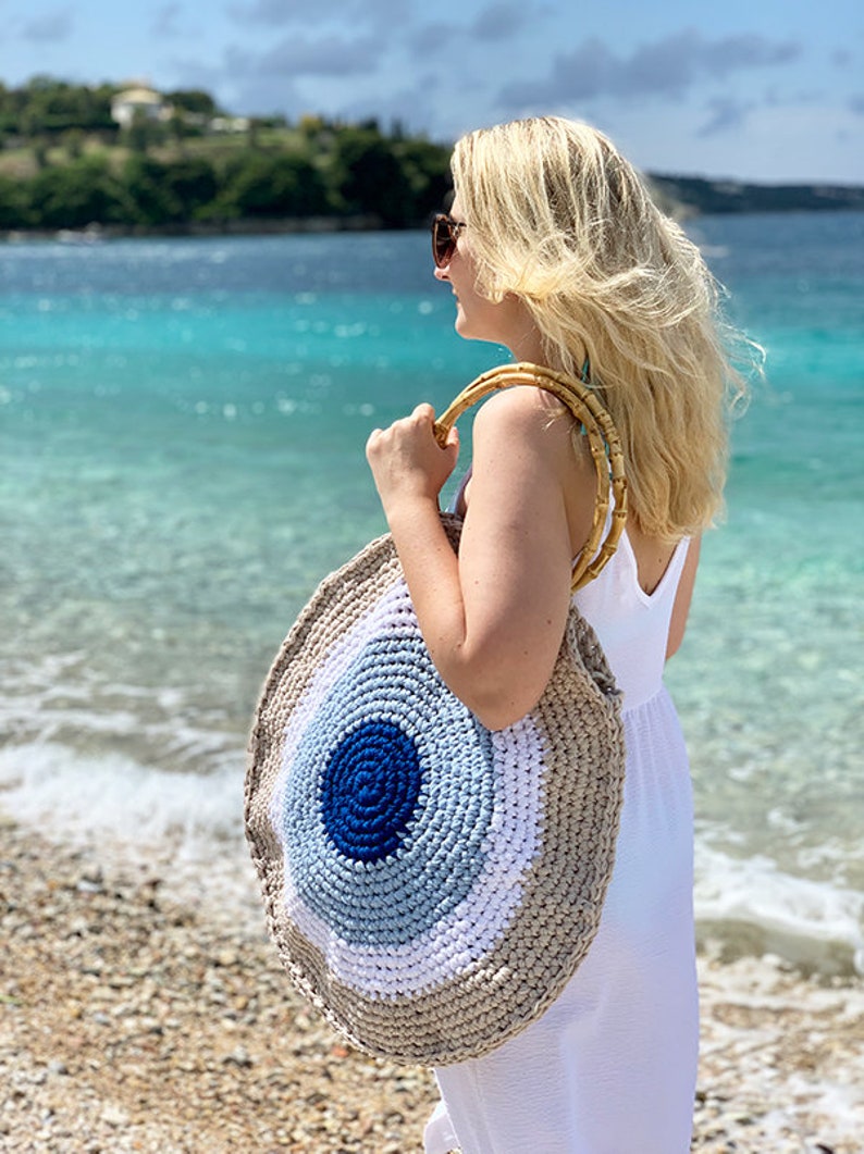 May include: A round, crocheted beach bag with a bamboo handle. The bag features concentric circles of color: dark blue, light blue, white, and beige. The bag is being held on a beach with turquoise water in the background.