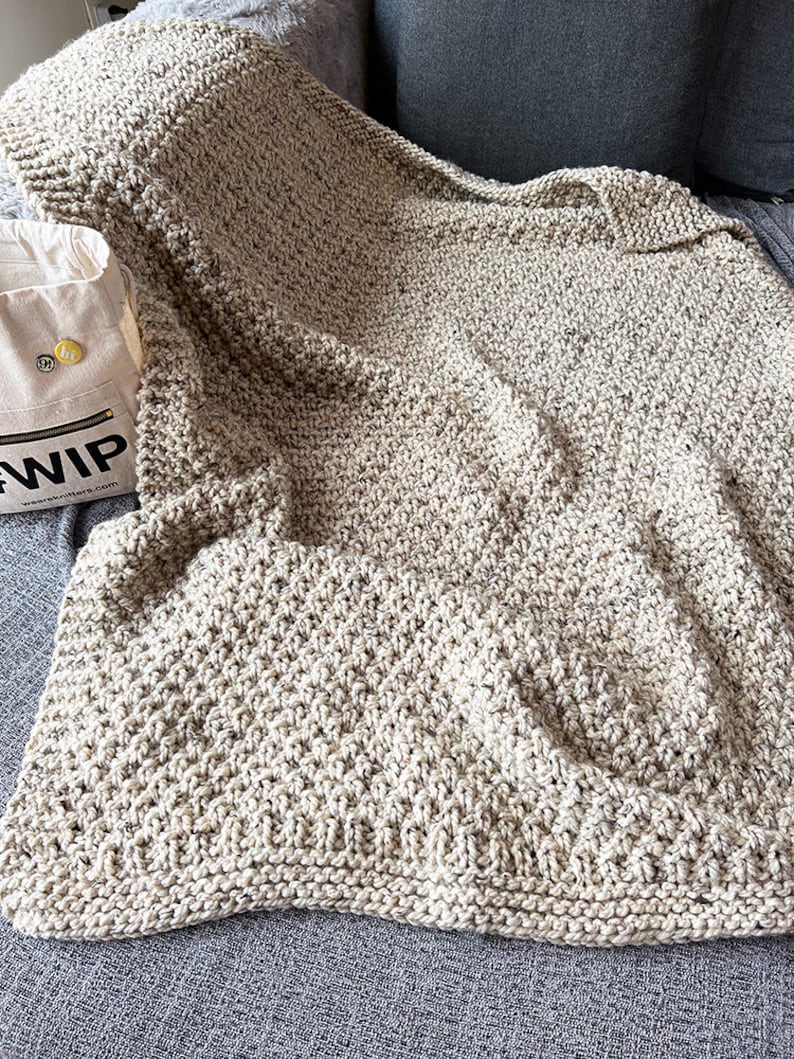 May include: A beige knitted throw blanket with a textured pattern. The blanket is folded and draped over a gray couch.