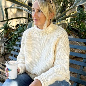 May include: A woman wearing a cream knitted jumper with a high neckline. The jumper has a textured stitch pattern and is made of a soft, cosy material.