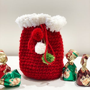 May include: A red crocheted drawstring bag with a white faux fur trim and green and white pom-poms. The bag is filled with wrapped chocolates.