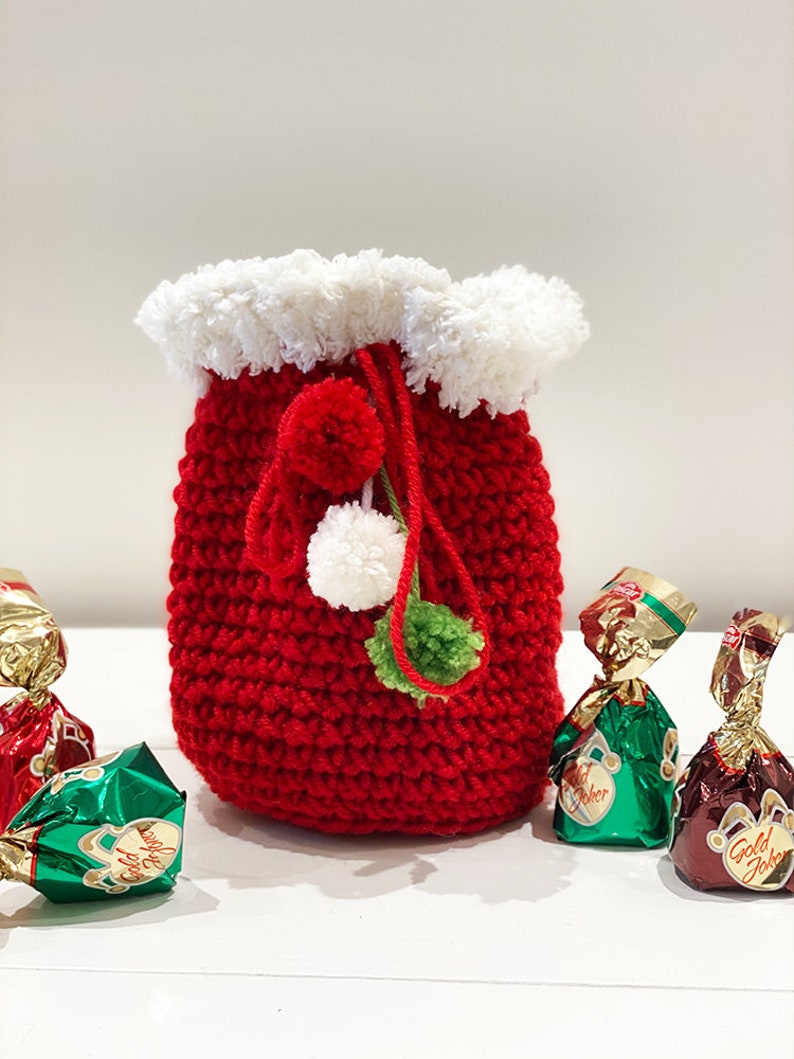 May include: A red crocheted drawstring bag with a white faux fur trim and green and white pom-poms. The bag is filled with wrapped chocolates.