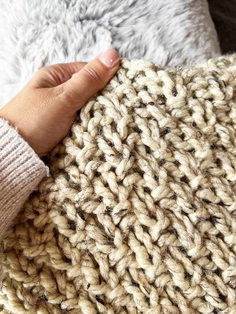 May include: Close-up of a chunky knit throw blanket in a cream color with a subtle flecked texture.