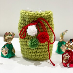 May include: A green crocheted gift bag with red and white pom poms. The bag is tied with a red ribbon and has a green pom pom on the side.