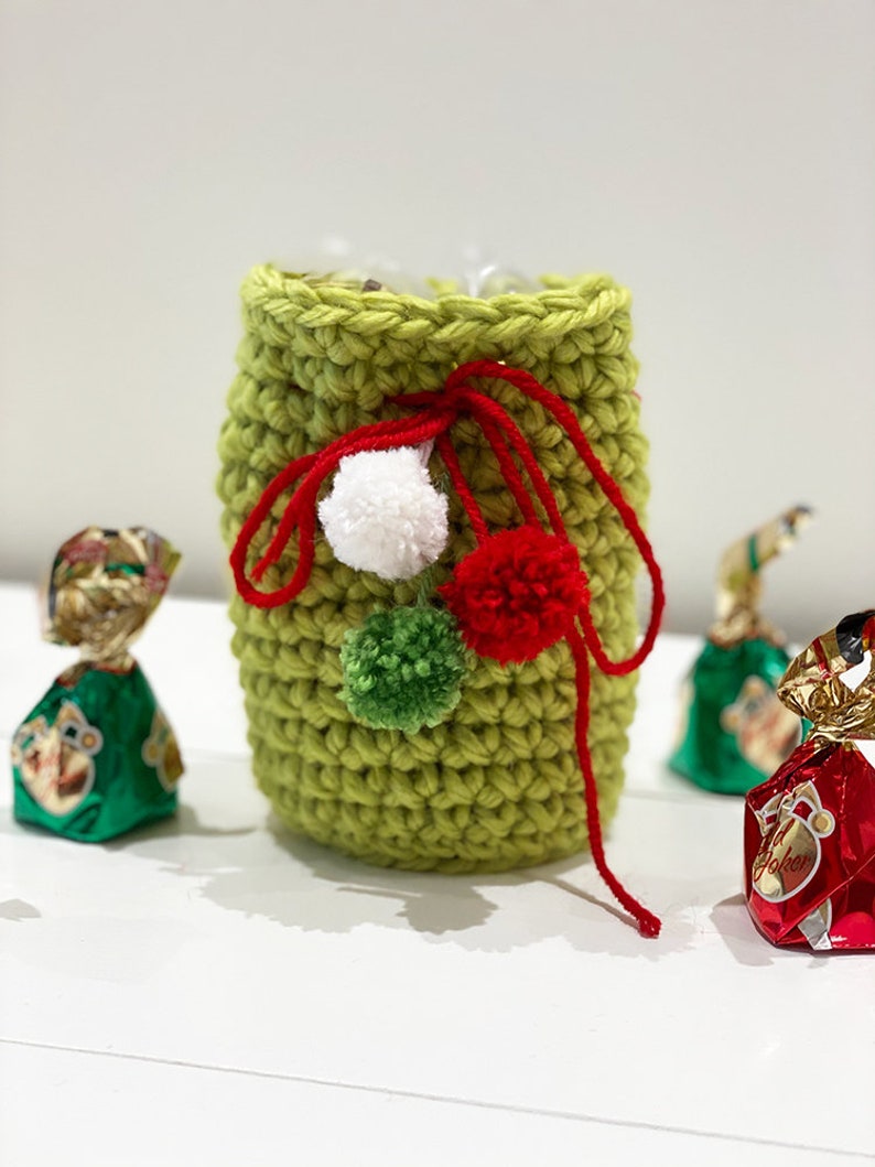 May include: A green crocheted gift bag with red and white pom poms. The bag is tied with a red ribbon and has a green pom pom on the side.