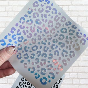 May include: A sheet of iridescent leopard print stencils. The stencils feature a repeating pattern of leopard spots in shades of blue, purple, and pink. The stencils are held up against a white brick wall.