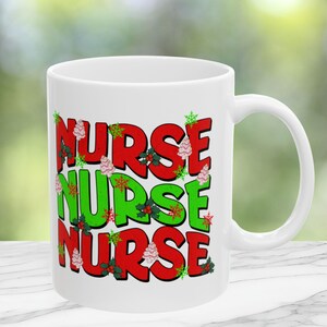 May include: White ceramic mug with the word "NURSE" stacked three times in red and green, decorated with snowflakes, Christmas trees, and holly. The mug has a white handle and is set against a blurred green background.