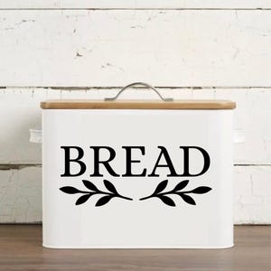 May include: White bread box with a wooden lid and a silver handle. The word "BREAD" is displayed in large, black letters, with decorative leaf accents. The box is rectangular and sits on a wooden surface.