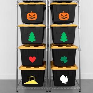 May include: Eight black plastic storage bins with yellow lids arranged on a metal wire shelving unit. Each bin features a distinct vinyl decal: an orange jack-o'-lantern, a green pine tree, a red heart, a green clover, three yellow crosses, and a white turkey.