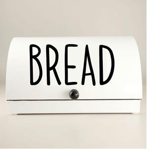 May include: A white bread box with a curved top and a black knob. The word "BREAD" is prominently displayed in large, black, bold letters across the front of the box. The box has a simple, modern design.