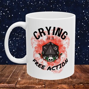 May include: White ceramic mug with the text "CRYING is a FREE ACTION" in black lettering. The design includes a black D20 die, flowers, and a skull on a red smoky background. The mug is on a wooden surface.