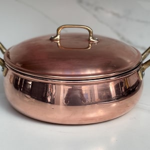 May include: A copper cooking pot with a lid and two side handles, and a handle on the lid. The pot has a shiny, reflective surface and brass-coloured handles. The pot is on a white surface.