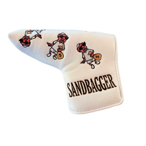 Puede incluir: Cubierta blanca para la cabeza del palo de golf con un patrón repetido de personajes de dibujos animados que sostienen bolsas de dinero. El texto "SANDBAGGER" está bordado en oro y negro en la parte inferior de la cubierta.