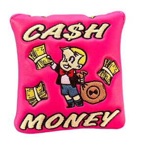 May include: Pink golf club head cover with an embroidered cartoon character holding a bag of money and surrounded by money. The text "CASH MONEY" is embroidered on the cover.