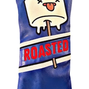 Könnte beinhalten: Blaue Golfschlägerkopf-Hülle mit einem weißen Marshmallow auf einem Stiel, der eine rote Zunge herausstreckt, und dem Text "ROASTED" auf einem blau-weißen Banner. Die Unterseite der Hülle trägt den Text "B&B GOLF" in Weiß.