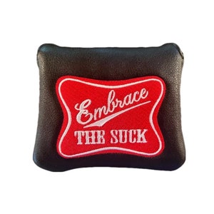 Puede incluir: Porta pelotas de golf de cuero negro con un parche rojo y blanco que dice "Embrace the Suck".
