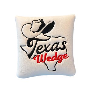 May include: Cream-coloured golf club head cover with a black and red embroidered design. The design features a cowboy hat, the word "Texas" in black script, and the word "Wedge" in red, within the outline of Texas.