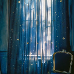 Pode incluir: Uma cadeira de veludo azul com moldura dourada fica em frente a uma janela com cortinas azuis adornadas com estrelas douradas. As cortinas s&atilde;o transparentes e permitem que a luz brilhe.