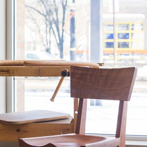 May include: A wooden chair with a curved back and seat, sitting in front of a wooden workbench with a vise. The chair and workbench are in a room with a window overlooking a snowy scene.