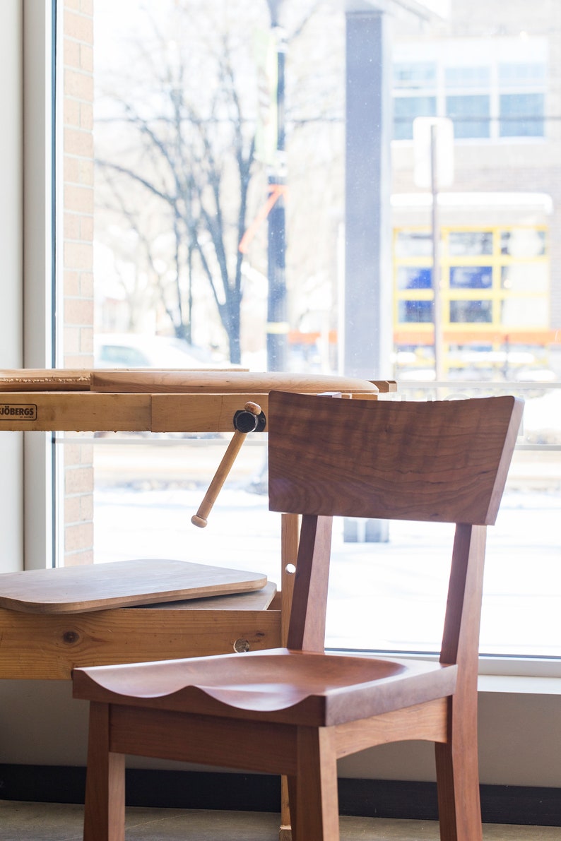 May include: A wooden chair with a curved back and seat, sitting in front of a wooden workbench with a vise. The chair and workbench are in a room with a window overlooking a snowy scene.