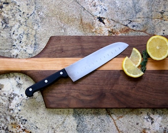 Puede incluir: Una tabla de cortar de madera con un diseño de nogal y arce. La tabla tiene un mango y un cuchillo apoyado en ella. Hay rodajas de limón en la tabla.