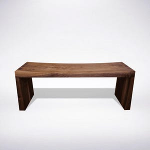 Peut inclure: Un banc rectangulaire en bois présentant un motif de grain de bois brun foncé naturel. Le banc possède une surface supérieure légèrement incurvée et deux pieds verticaux solides en forme de dalle de chaque côté, formant une base stable.