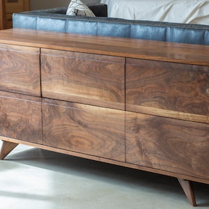 May include: A long, rectangular wooden dresser with six drawers. The dresser is made of dark wood with a natural grain pattern. It has a modern design with angled legs.