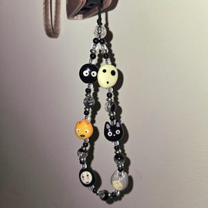May include: A beaded phone charm featuring cartoon character beads. The charm includes black and white beads, a black cat, a round orange character, and a gray character. The charm is attached to a black cord.