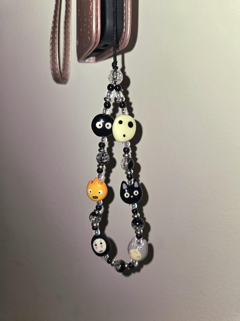 May include: A beaded phone charm featuring cartoon character beads. The charm includes black and white beads, a black cat, a round orange character, and a gray character. The charm is attached to a black cord.