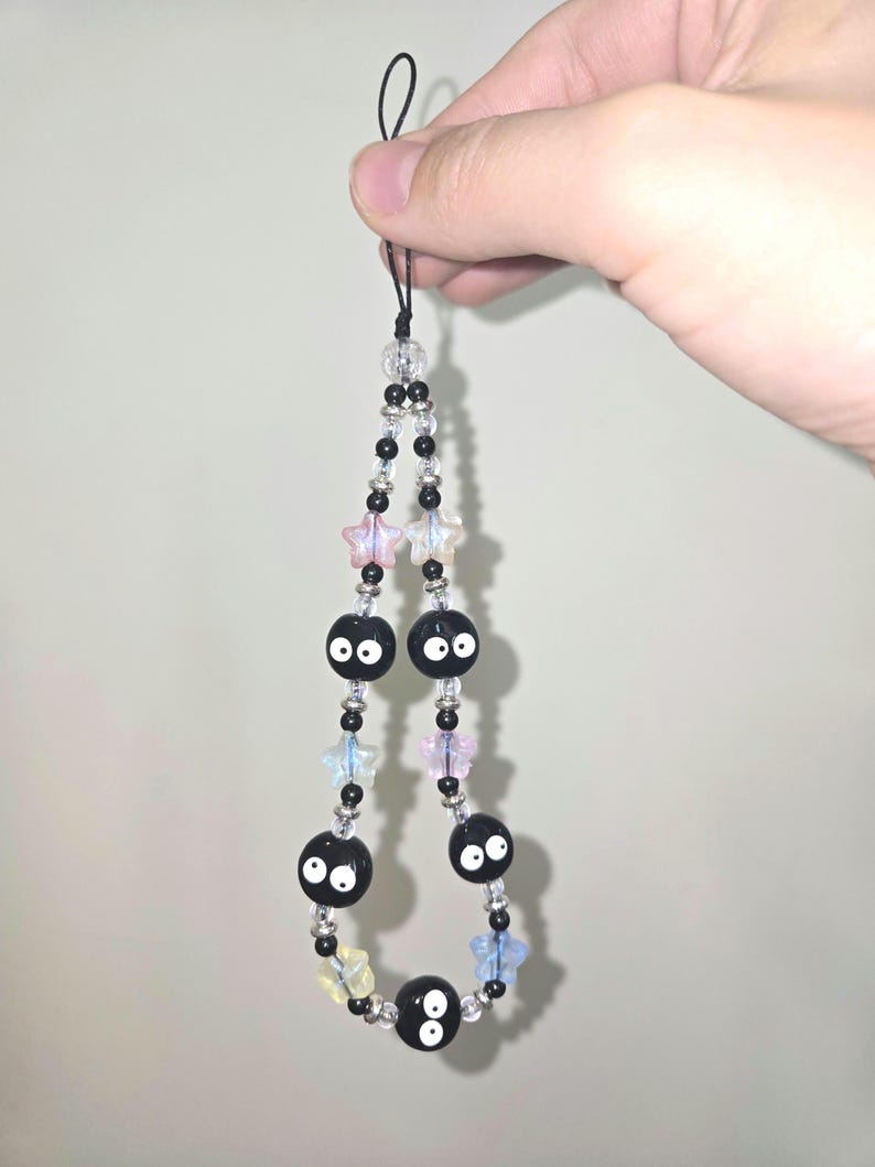 May include: Black and white beaded phone charm with a string loop at the top. The beads are shaped like stars and have a black bead with two white eyes in the center.