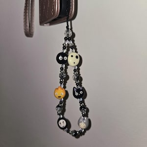 May include: A black beaded phone charm featuring cartoon character beads. The charm includes a variety of character faces in black, white, gray, and orange. The charm is attached to a rose gold phone case.
