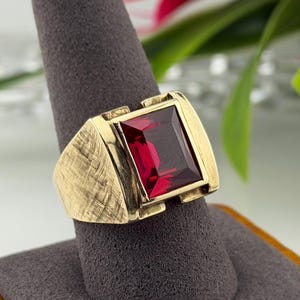 May include: A gold-toned ring with a large, rectangular, deep red gemstone. The ring has a textured band and a rectangular setting that frames the stone. The ring is displayed on a gray ring holder.