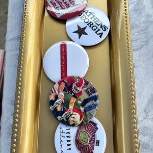 Puede incluir: Una colección de botones redondos con varios diseños y texto. Un botón dice "THE BEST GIRLS ARE GEORGIA GIRLS". Otro dice "ATHENS GEORGIA" con una estrella. Un tercero dice "sorry i can't its game day". Los botones están en una bandeja dorada.